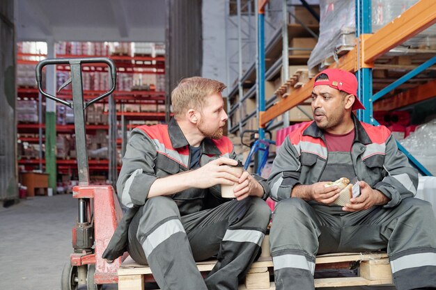 Foto due giovani lavoratori interculturali seduti su un carrello industriale in un ambiente di magazzino e discutono dopo i piani di lavoro durante la pausa pranzo