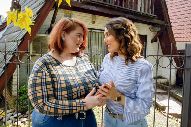 Two young latin friends standing talking to each other and using the phone outside at noon