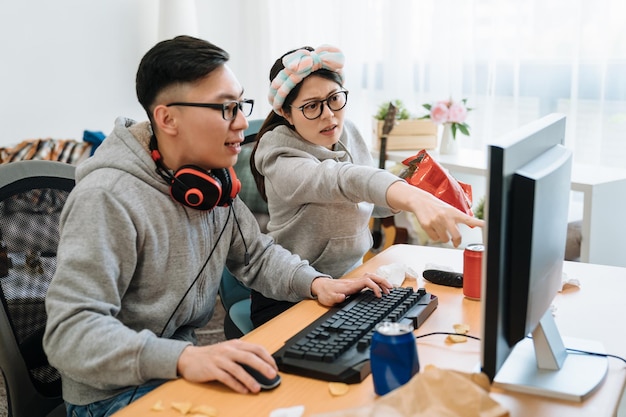 two young lazy asian couple having fun playing online video game at home. girlfriend holding pack of chips and point to computer screen teaching her boyfriend. man in headphones control on keyboard