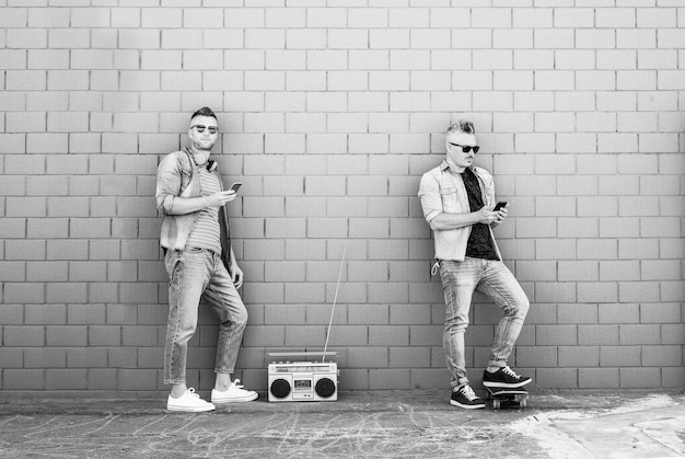 Two young man looking at mobile phone lean against brick wall Couple of cool guys listening music with vintage boombox stereo outdoor in the street Concept of tech social network and music trends