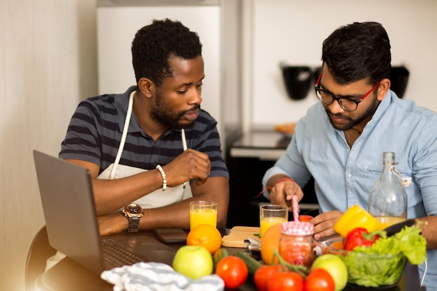 Due giovani uomini in abiti casual seduti a tavola, preparando insalata, tagliando verdure, usando il computer portatile in cucina al mattino. cibo sano, cucina, concetto di tecnologia.