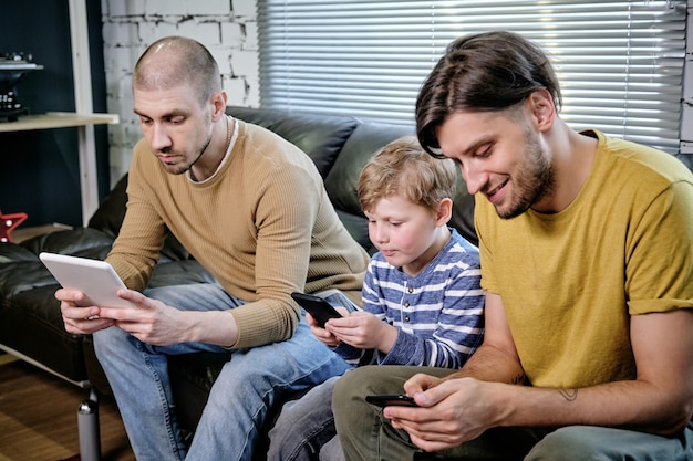 Two young men and little boy using mobile gadgets when sitting on sofa at home