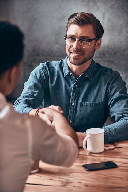 Two young men in smart casual wear shaking hands while sitting at the office desk