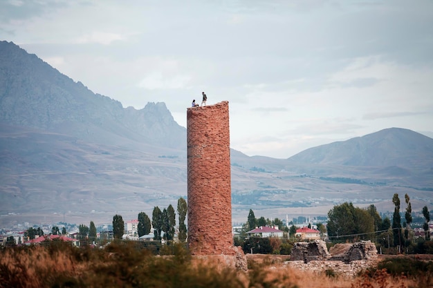 Two young men stand on top of the ruined of the medieval Osman Mosque and its minaret in the Old City of Van against the backdrop of mountains, near the famous Van Fortress, Van, Turkey.