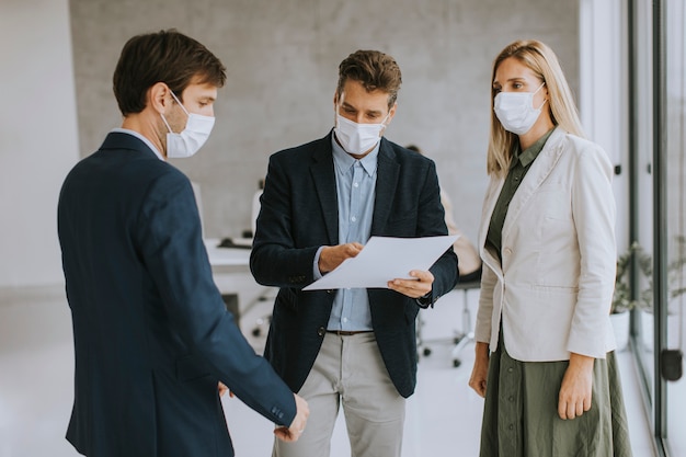 Two young men and woman discussing with paper in hands indoors in theoffice with young people works behind them