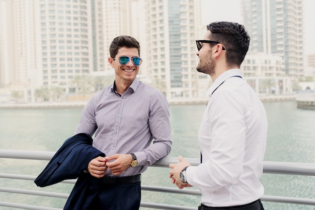 Two young modern businessman in Dubai Marine leaned on fence. Standing and discussing about business.