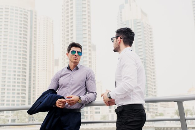 Two young modern businessman in Dubai Marine leaned on fence. Standing and discussing about business.