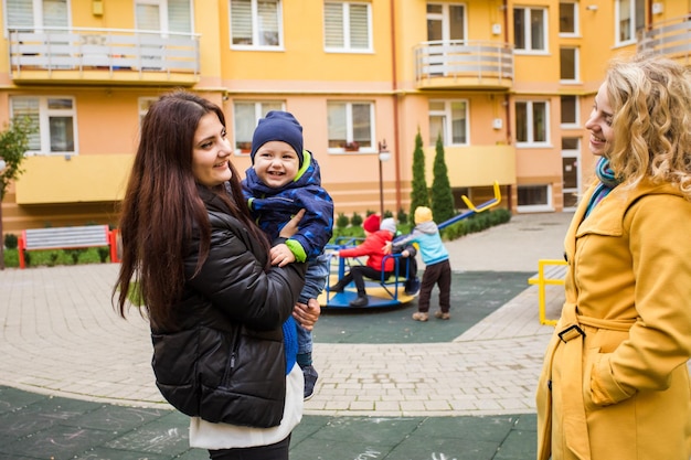 遊び場の前で屋外で話している2人の若い母親友達と話している小さな男の子を抱いている金髪の女性ニュースと経験を共有する