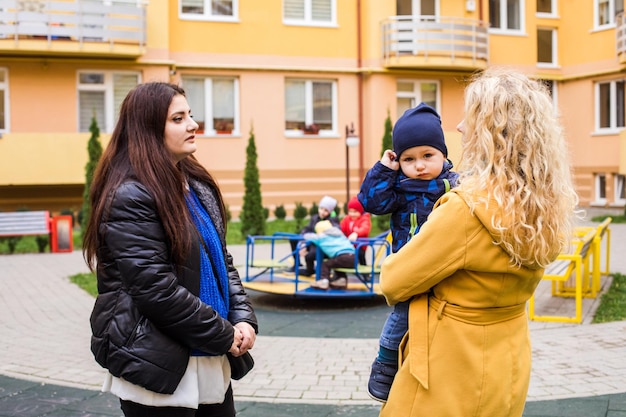 Two young mothers talking outdoors in front of playground Blonde woman holding little boy talking to her friend Sharing news and experience