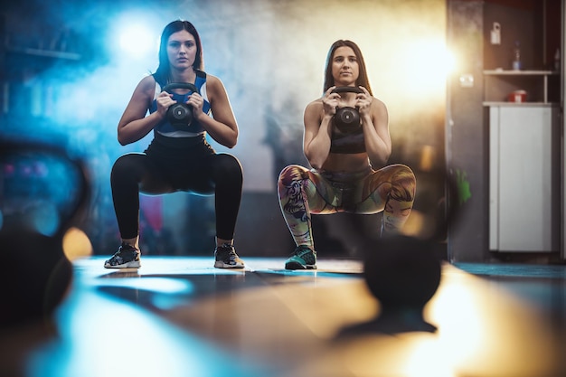 Foto due giovani donne muscolose e carine stanno facendo squat con kettlebell in palestra.