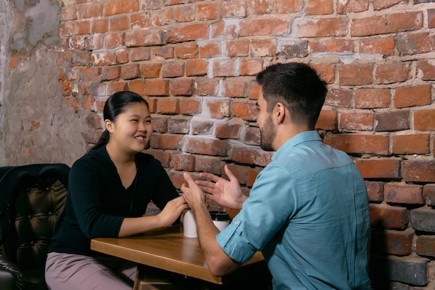 Two young people a guy and a girl talking about something fun while sitting at a table in a cafe on the background of a rough brick wall