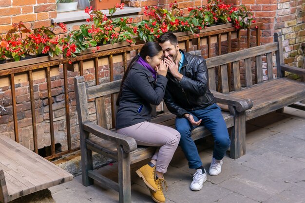 Two young people a guy and a girl whisper about something and laugh sitting on a bench outdoors