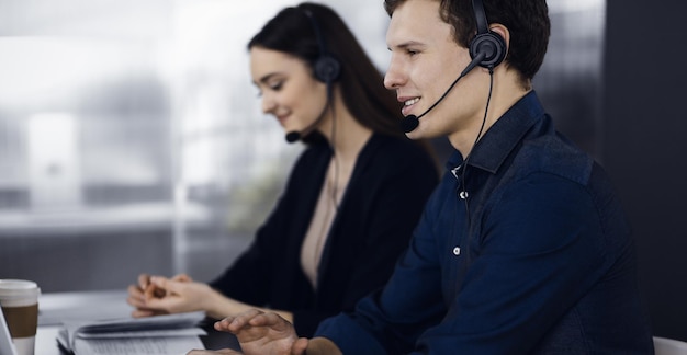 Two young people in headsets are talking to the clients, while sitting at the desk in a modern office. Focus on man in a blue shirt. Call center operators at work.