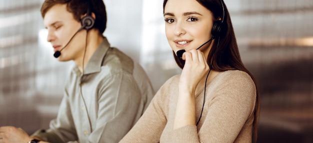 Two young people in headsets are talking to the clients, while sitting at the desk in an office. Focus on woman. Call center operators at work.
