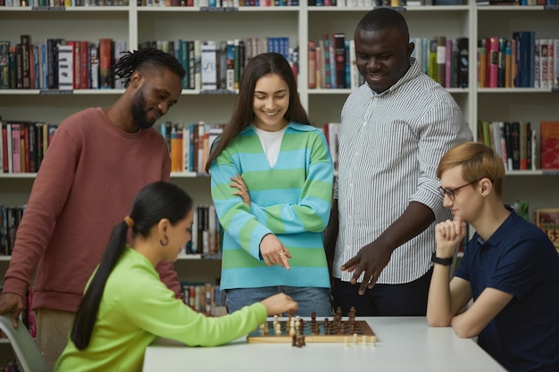 Two young people playing chess with group of friends watching and cheering