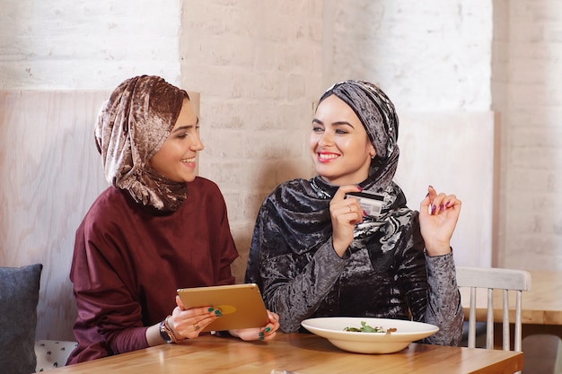 두 명의 젊은 예쁜 무슬림 여성이 전자 태블릿을 사용하여 온라인으로 이야기하고 쇼핑합니다.