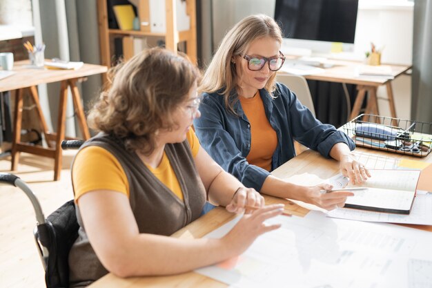 Two young pretty office managers sitting by table and looking at plan of work on page of notebook during discussion of its points at meeting