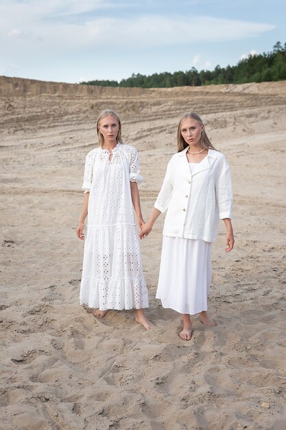 Two young pretty twins with long blond hair posing at sand quarry in elegant white dresses