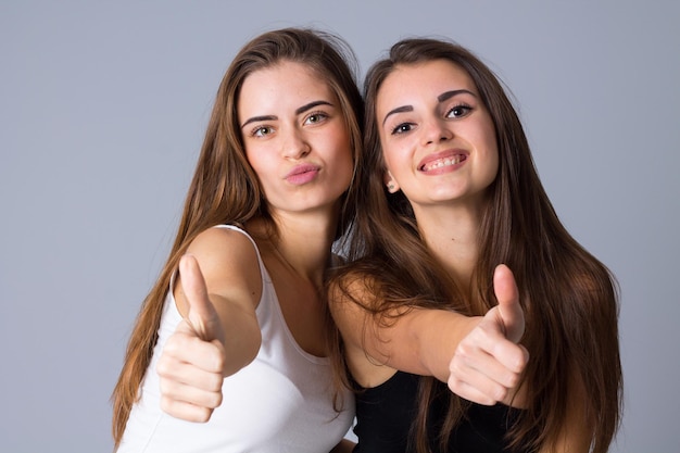 Two young pretty women in black and white shirts showing thumbs up on gray background in studio