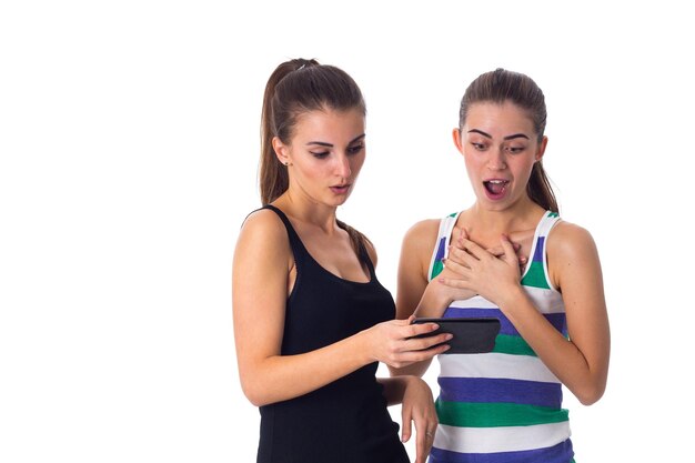 Two young pretty women in striped and black shirts using smartphone on white background in studio