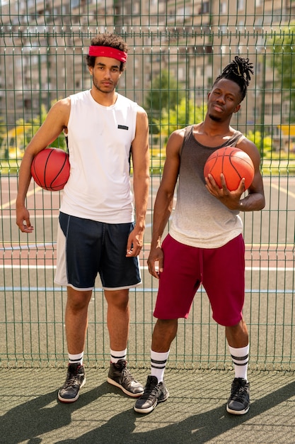 Two young professional multicultural basketball players standing on playground by fence on summer day