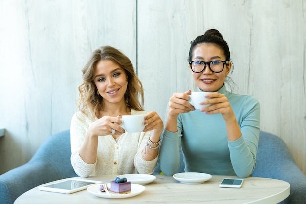 写真 カフェのテーブルのそばの快適なソファに座っているカプチーノのカップを持つ2人の若い安らかな異文化間の女性