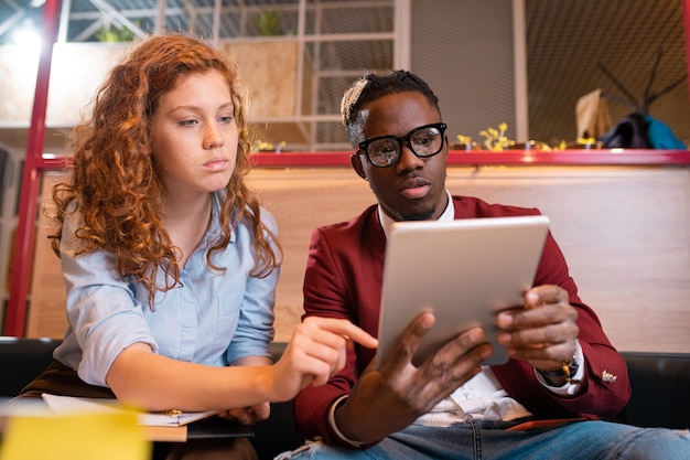 Photo two young serious intercultural employees in casualwear discussing electronic document on touchpad display at meeting