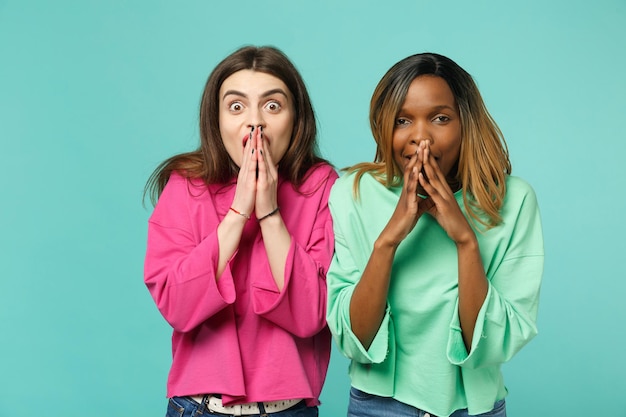 Two young shocked surprised women friends european and african american in pink green clothes isolated on blue turquoise wall background, studio portrait. People lifestyle concept. Mock up copy space.