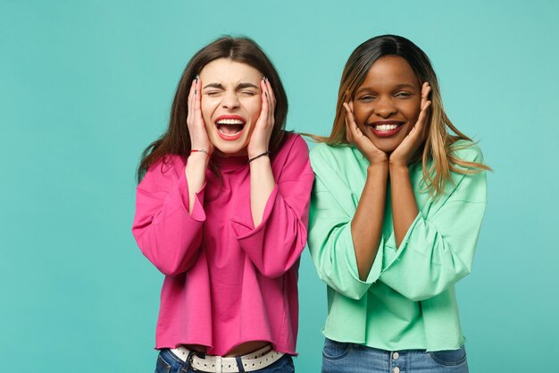 Two young shocked surprised women friends european and african american in pink green clothes isolated on blue turquoise wall background, studio portrait. People lifestyle concept. Mock up copy space.