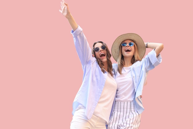 Две молодые улыбающиеся хипстерские женщины в летней одежде позируют на розовом фоне Женщина показывает положительные эмоции на лице
