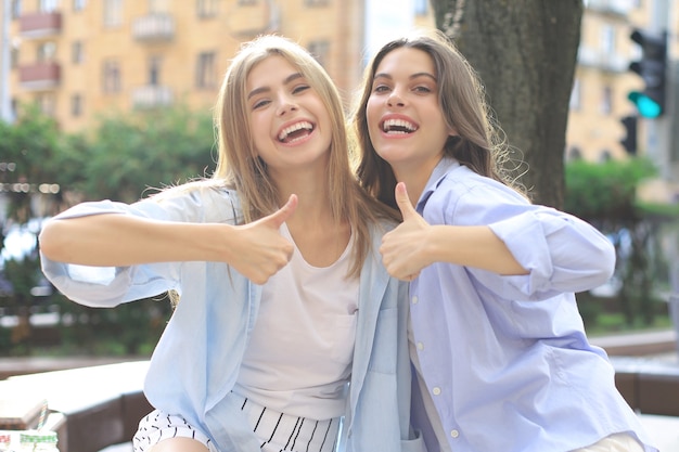 Two young smiling hipster women in summer clothes posing on street.Female showing positive face emotions.