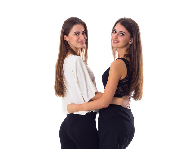 Two young smiling women in white and black shirts hugging on white background in studio