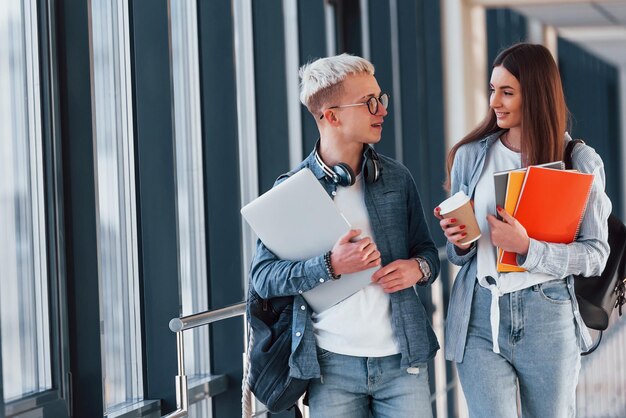 Two young student friends together in a corridor of a college walking together and holding notepad laptop and cup of drink