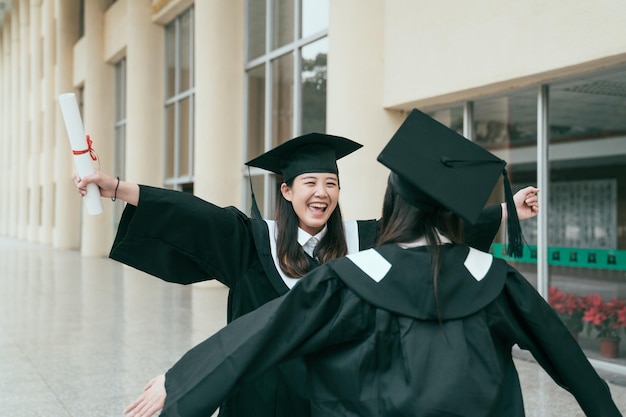 Two young student girls dressed in black graduation gown\
smiling holding diplomas hugging. woman open arms outstretched\
giving love to best friend on finished university last day in\
building hall.