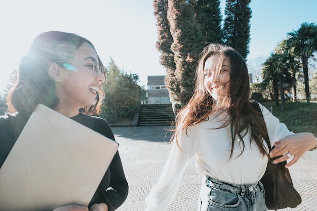 Two young student woman going to class while talking and\
laughing outdoors the faculty during a bright day. university\
studies, college papers, modern style. preparing a presentation for\
class.