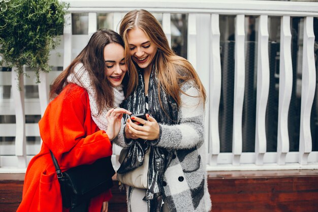 2つの若いスタイリッシュな女性は、秋の都市に立って、電話を使用して