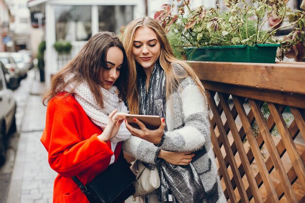 2つの若いスタイリッシュな女性は、秋の都市に立って、タブレットを使用して