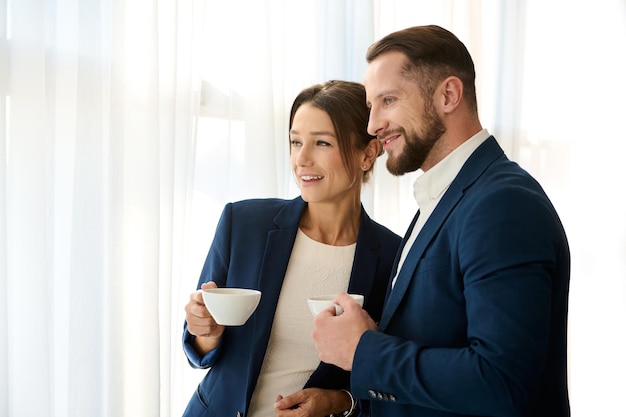 Two young successful handsome business partners standing next to each other with a cup of coffee in their hands and smiling, confidently looking out the window. Copy space for ad