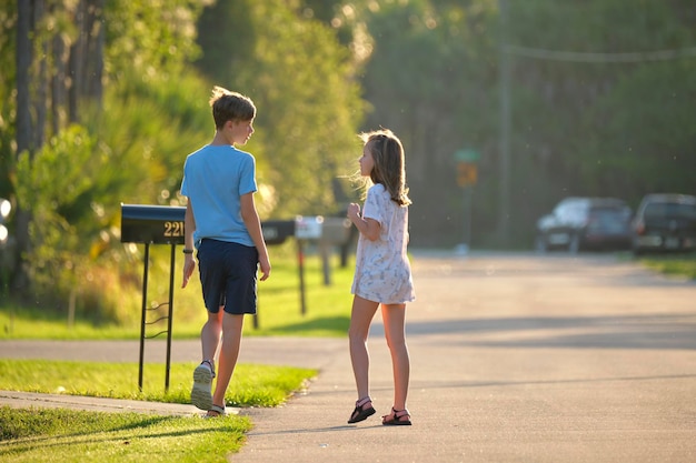 Photo two young teenage children girl and boy standing and talking together outdoors on bright sunny day on rural street side