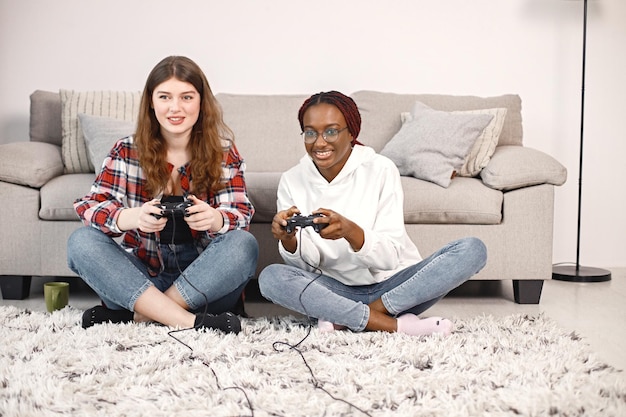 Две молодые девочки-подростки сидят на полу возле кровати и играют в PlayStation.
