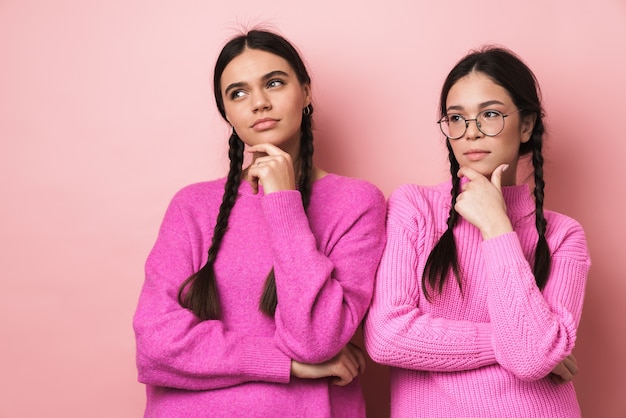 two young teenage girls with braids in casual clothes touching chins and looking aside isolated over pink wall