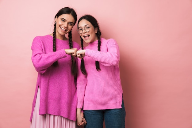 two young teenage girls with braids smiling and bumping their fists together isolated over pink wall