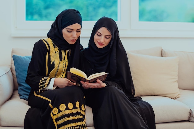 two young traditional muslim women read quran on the sofa before iftar dinner during a ramadan feast at home.