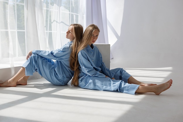 Две молодые девушки-близнецы в одинаковых синих костюмах сидят на полу с белым циклорамой