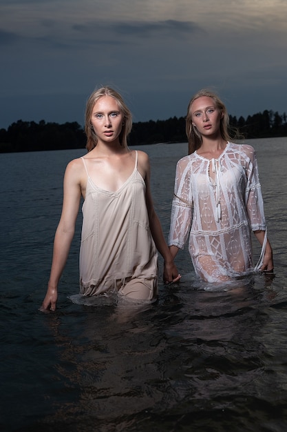 긴 금발 머리가 여름 밤에 호수의 물에 가벼운 드레스를 입고 포즈를 취하는 두 젊은 쌍둥이 자매, 카메라를보고