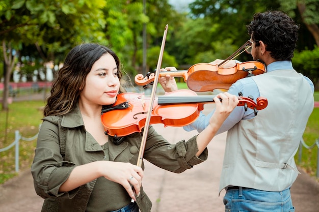 Two young violinists standing playing violin in a park Portrait of man and woman together playing violin in park Violinist man and woman back to back playing violin in a park outdoors