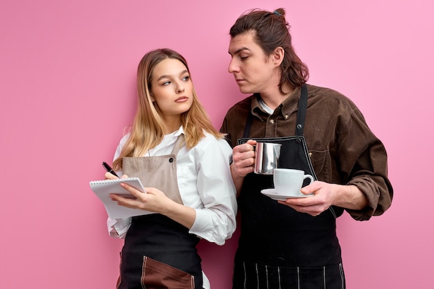 two young waiters in apron discuss orders, ready to serve clients, friendly waitress stand writing making notes