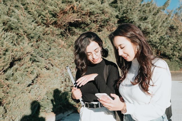 Две молодые женщины-студентки тихо разговаривают во время занятий в колледже в солнечный день, показывая что-то по телефону, весело проводя время Вернувшись в школу концепция Мультикультурные студенты в университете