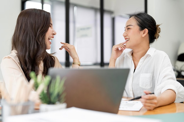 Две молодые женщины, работающие вместе, улыбаются и смеются от счастья в офисе