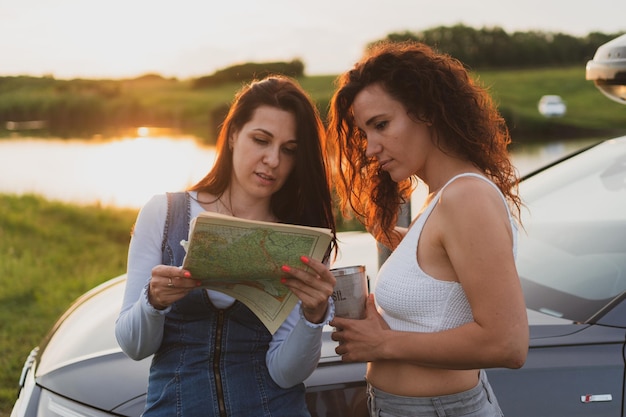 Two young women are traveling on the roads by car stopped at\
the side of the road and look at the map drinking coffee from a\
thermos vacation concept